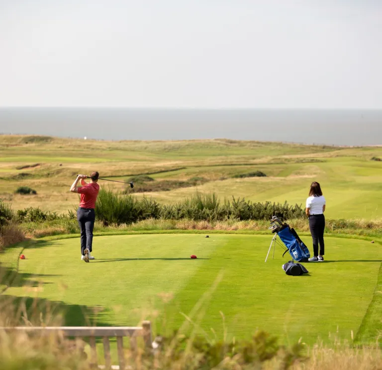Mae dau golffwr yn sefyll ar grewn, mae'r un ar y chwith yn cymryd y swing. Gellir gweld y môr y tu hwnt i grawn y ffordd.