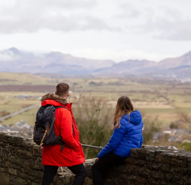dau unigolyn, un mewn côt law goch, a’r llall mewn côt law las, yn sefyll wrth hen wal gerrig yn edrych ar olygfa o gaeau gwyrdd a bryniau, a’r awyr yn niwlog