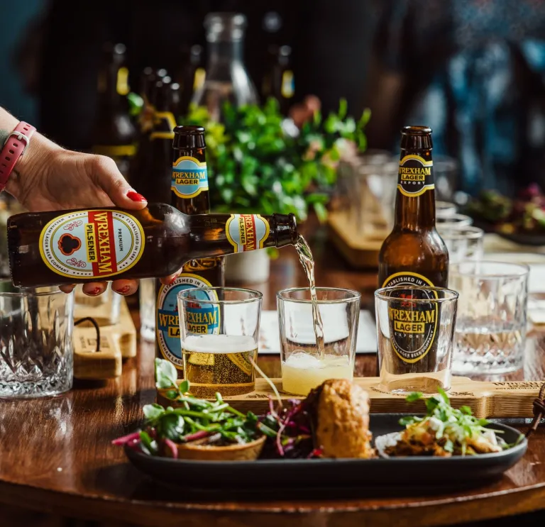 Platiad o fwys setil tapas a photel brown o Wrexham Lager yn cael ei roi mewn gwydr. 