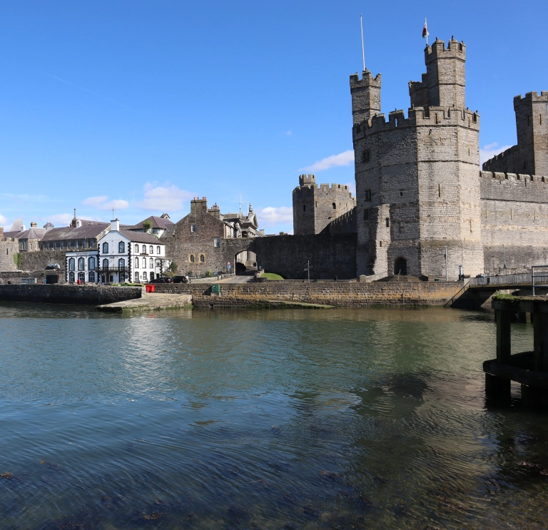 Castell Caernarfon wrth lan y dŵr, gyda’i furiau cerrig mawreddog yn cael eu hadlewyrchu yn nyfroedd tawel yr harbwr o dan awyr las.