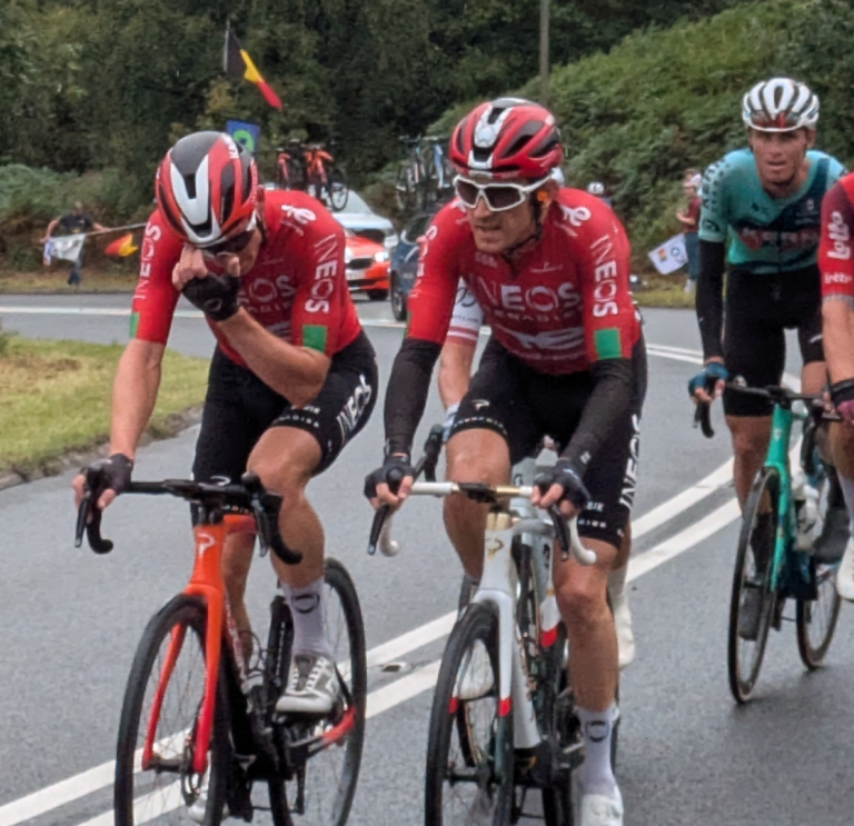 Grŵp o feicwyr ffordd proffesiynol mewn citiau rasio yn reidio'n agos gyda'i gilydd, gyda Geraint Thomas ar y blaen, ar ffordd wledig yn ystod digwyddiad beicio Tour of Britain 2025.