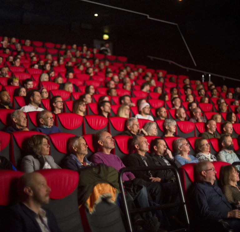 Cynulleidfa yn eistedd mewn cadeiriau coch y tu mewn i awditoriwm sinema.