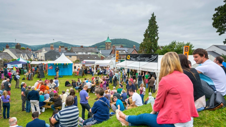 Pobl yn eistedd ar y gwair ar fryncyn yn edrych i lawr ar stondinau'r ŵyl fwyd