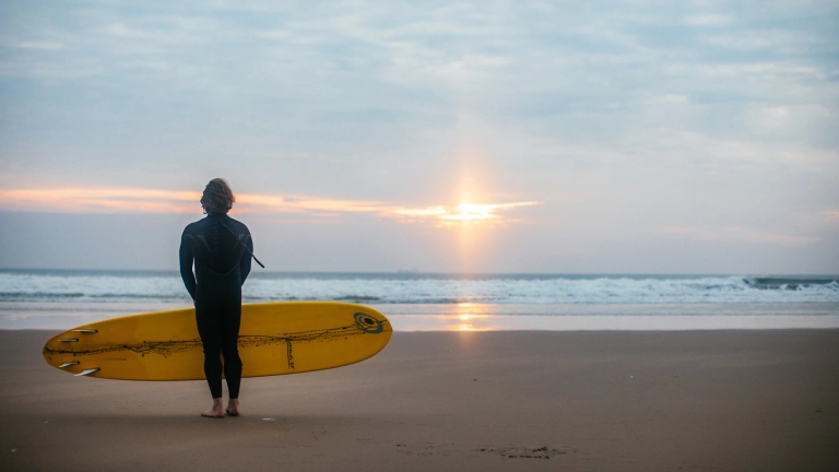 Dyn yn sefyll ar y traeth gyda'i gefn at y camera, yn wynebu'r môr ac yn dal bwrdd syrffio melyn