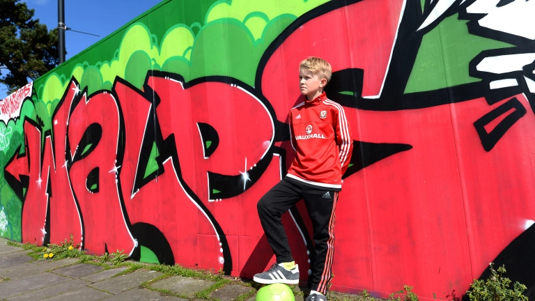 Bachgen yn pwyso ar wal gyda graffiti 'Cymru' arno, gydag un droed ar bêl-droed