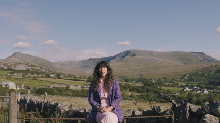 Landscape shot of singer Lisa Jen sat on a fence with the valley and surrounding mountains of Bethesda in the background.