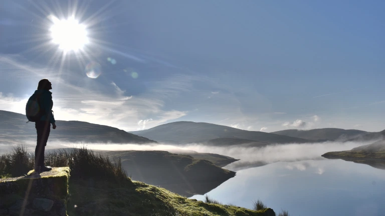 Rhywun yn edrych dros y mynyddoedd a llyn yn gynnar yn y bore.
