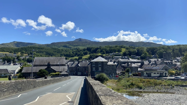Edrych tuag at Ddolgellau o dop y Bont Fawr. Mae'r tywydd yn braf ac mae modd gweld adeiladau creigiog y dref tu hwnt i'r bont. Yn y cefndir mae mynyddoedd gwyrdd.