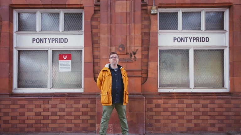 Dyn (Gareth Potter) mewn cot felen yn sefyll ar blatfform drên gydag arwydd Pontypridd.
