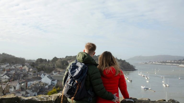dyn a dynes yn sefyll mewn castell gyda golygfa o’r môr a chychod.