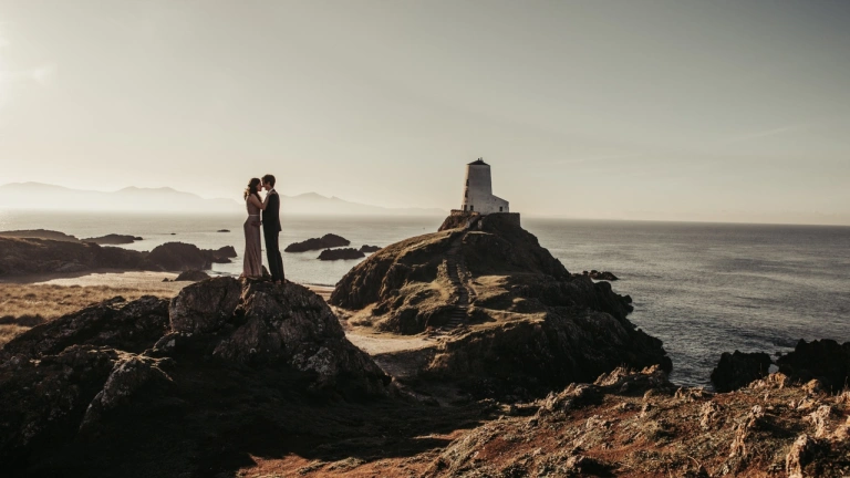 Llun priodas o gwpl yn cusanu ar ben craig ar draeth ynys Llanddwyn. Mae goleudy wen yn y cefndir.