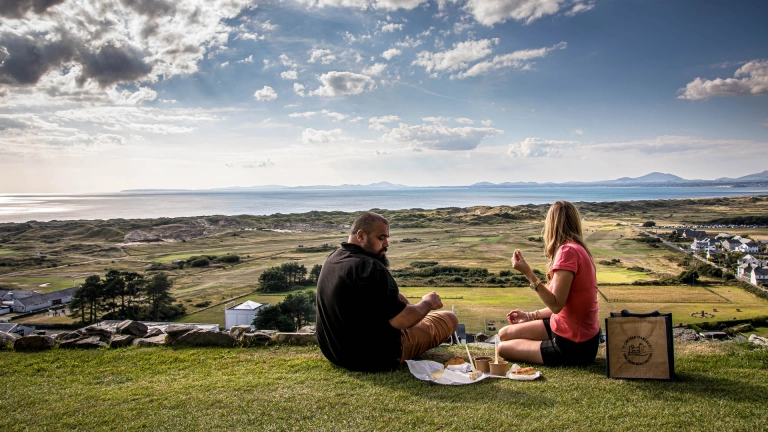 Dau berson yn cael picnic yn edrych dros cwrs golff.