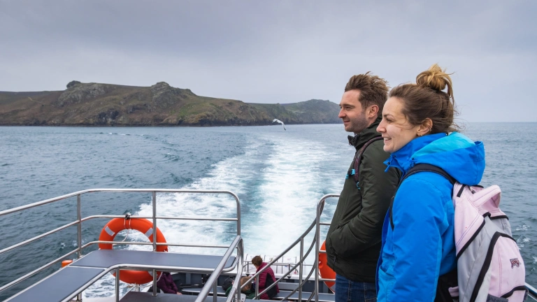 Dau berson ar gwch yn y môr, yn edrych ar ynys.