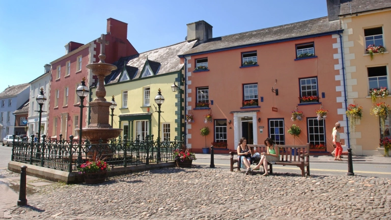 Sgwâr y dref, Llanymddyfri. Mae tai mawr crand oren a melyn ar y sgwrs, a dwy ddynes yn eistedd ar fainc.
