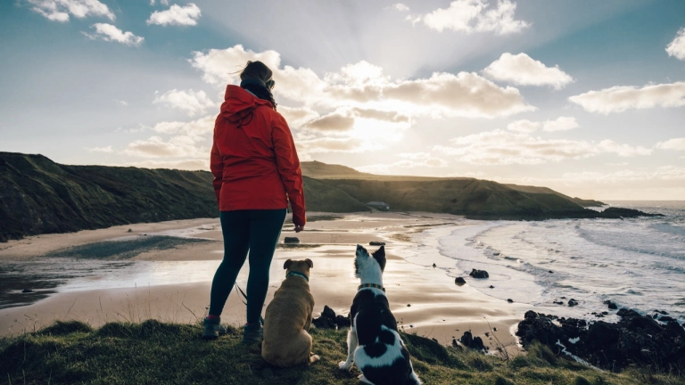Person mewn siaced goch yn sefyll gyda dau gi yn edrych allan ar fae tywodlyd.