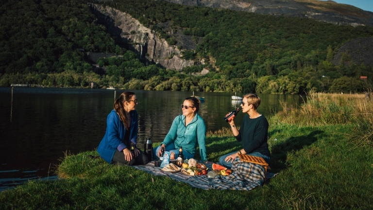 Tair menyw yn cael picnic ar lan llyn.