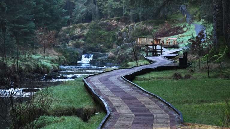 Llwybr eang mewn coetir sy'n arwain at bwynt gwylio, ochr yn ochr â'r afon