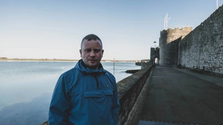 Person mewn cot las yn sefyll ger dŵr a muriau hynafol tref. 