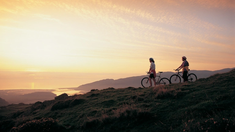Dau berson gyda beic mynydd yn edrych ar yr haul yn machlyd yn  aber afon Mawddach eastuary.