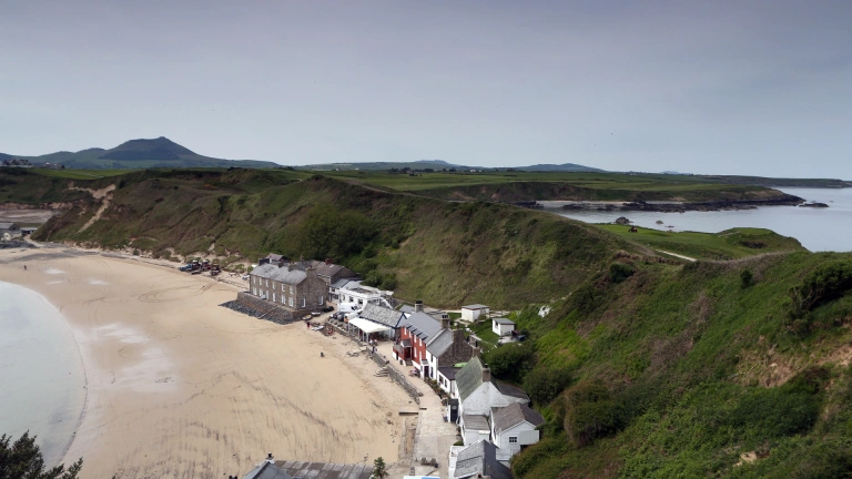 Llun o adeiladau ar y traeth, gyda bryniau gwyrdd y tu ôl.