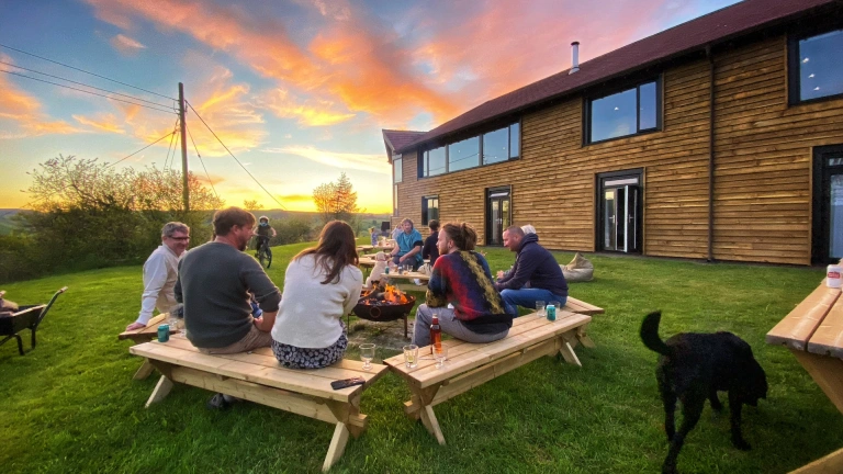 Grŵp o amgylch pwll tân y tu allan i adeilad arddull caban ar fachlud haul. 