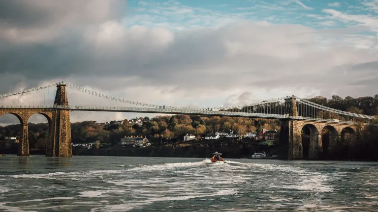 Llun o'r bont grog gyda choed a thai yn y cefndir a chwch ar Afon Menai