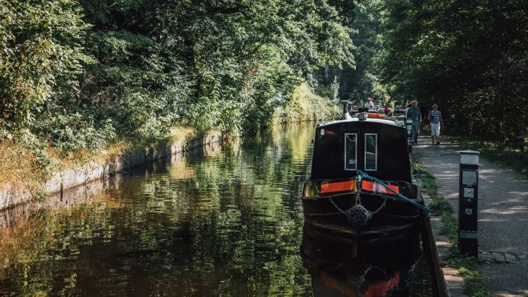 Black and red narrowboat moored on a tranquil canal lined with green trees, with people walking along the adjacent paved path.