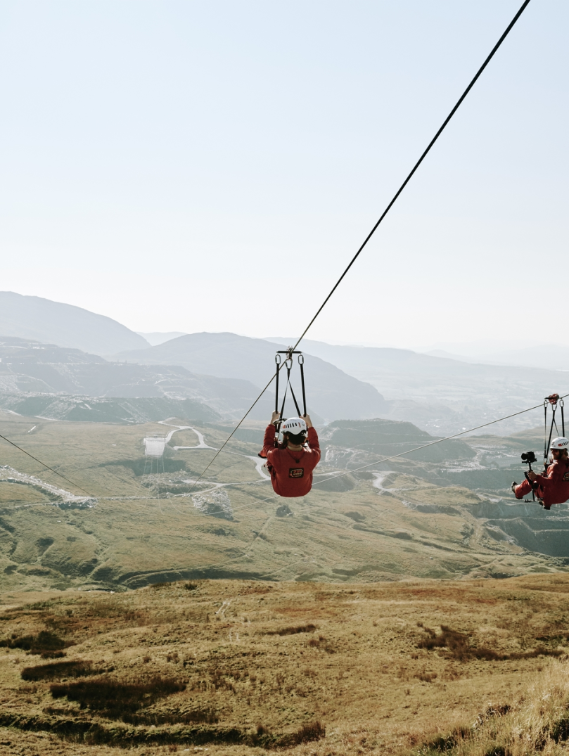 Tri o bobl yn marchogaeth zip-line yn uchel uwchben chwarel lechi.