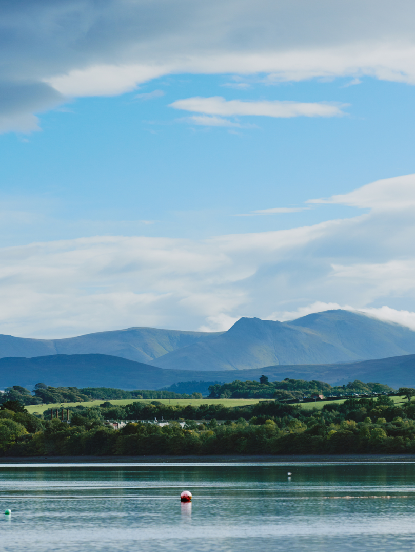 Llun o'r môr gyda choed a bryniau yn y pellter.