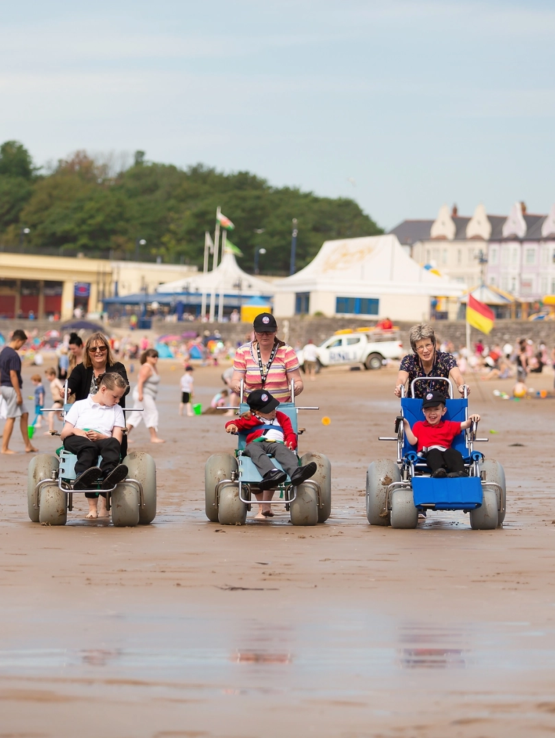 Tri pherson yn defnyddio cadeiriau olwyn traeth ar y traeth.