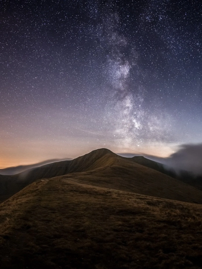 Awyr dywyll yn llawn sêr uwchben tirwedd fynyddig Bannau Brycheiniog gyda golau'r haul yn machlud ar y gorwel.