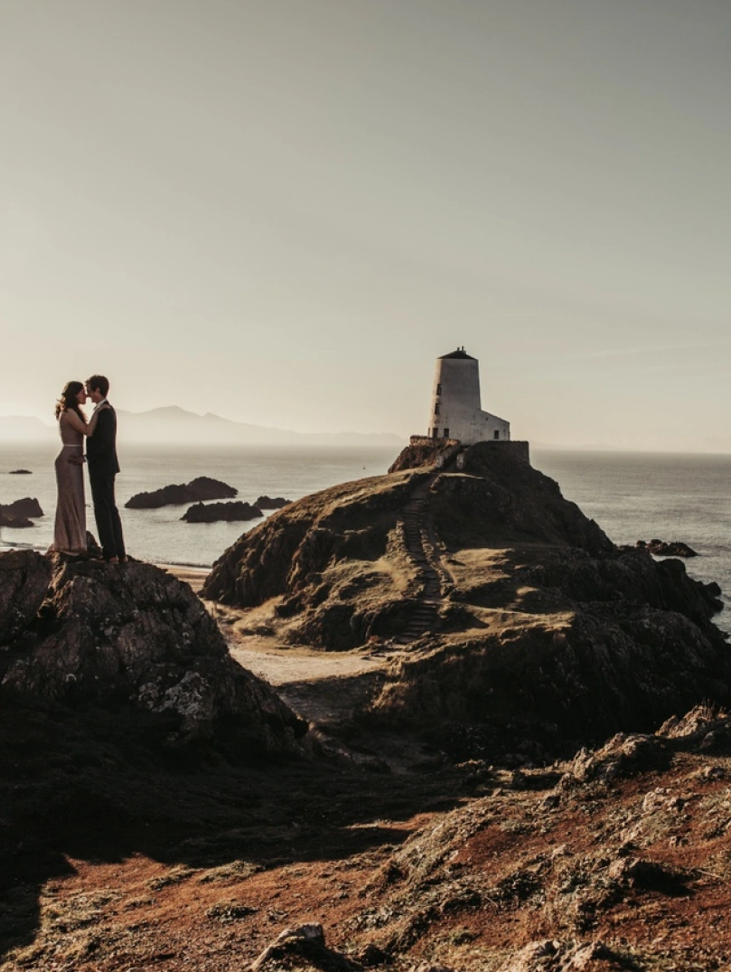 Llun priodas o gwpl yn cusanu ar ben craig ar draeth ynys Llanddwyn. Mae goleudy wen yn y cefndir.