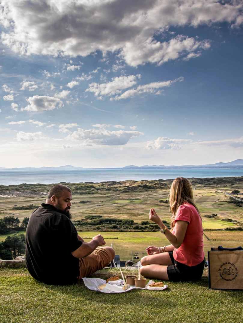 Dau berson yn cael picnic yn edrych dros cwrs golff.