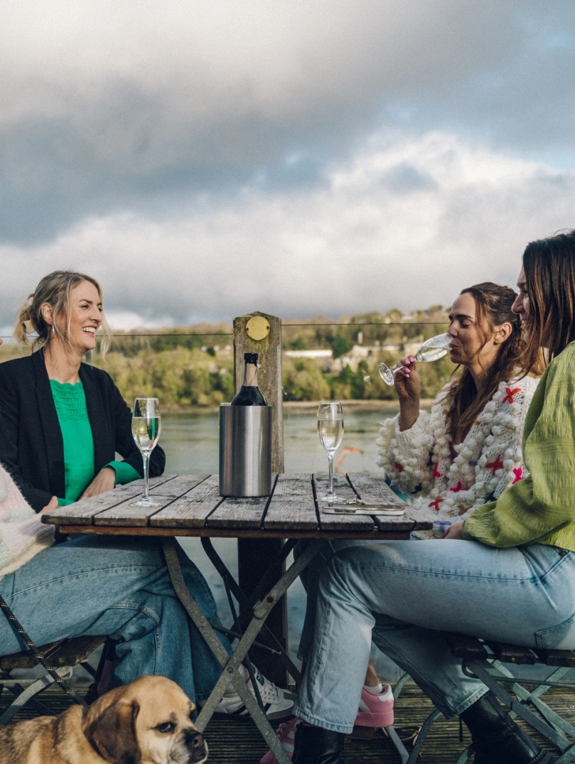 grŵp o fenywod yn yfed wrth fwrdd y tu allan gyda chi wrth eu traed a golygfeydd o Fenai.