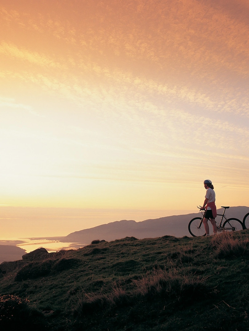 Dau berson gyda beic mynydd yn edrych ar yr haul yn machlyd yn aber afon Mawddach eastuary.
