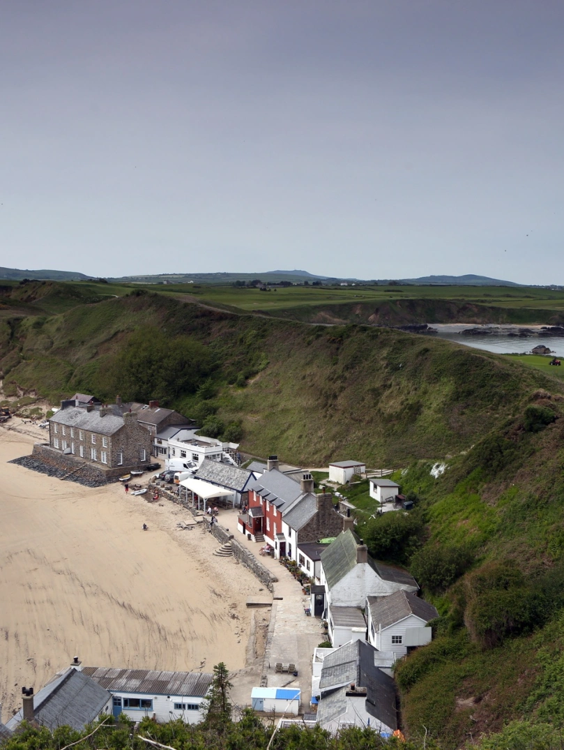 Llun o adeiladau ar y traeth, gyda bryniau gwyrdd y tu ôl.