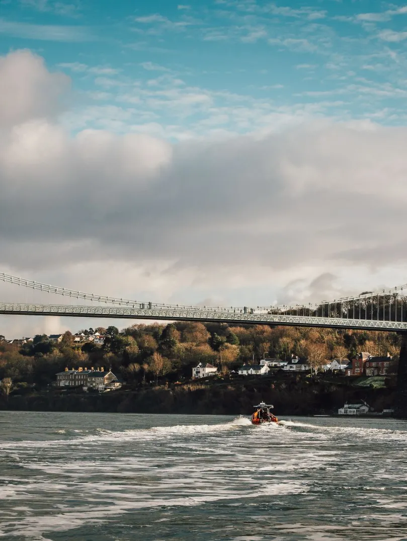 Llun o'r bont grog gyda choed a thai yn y cefndir a chwch ar Afon Menai