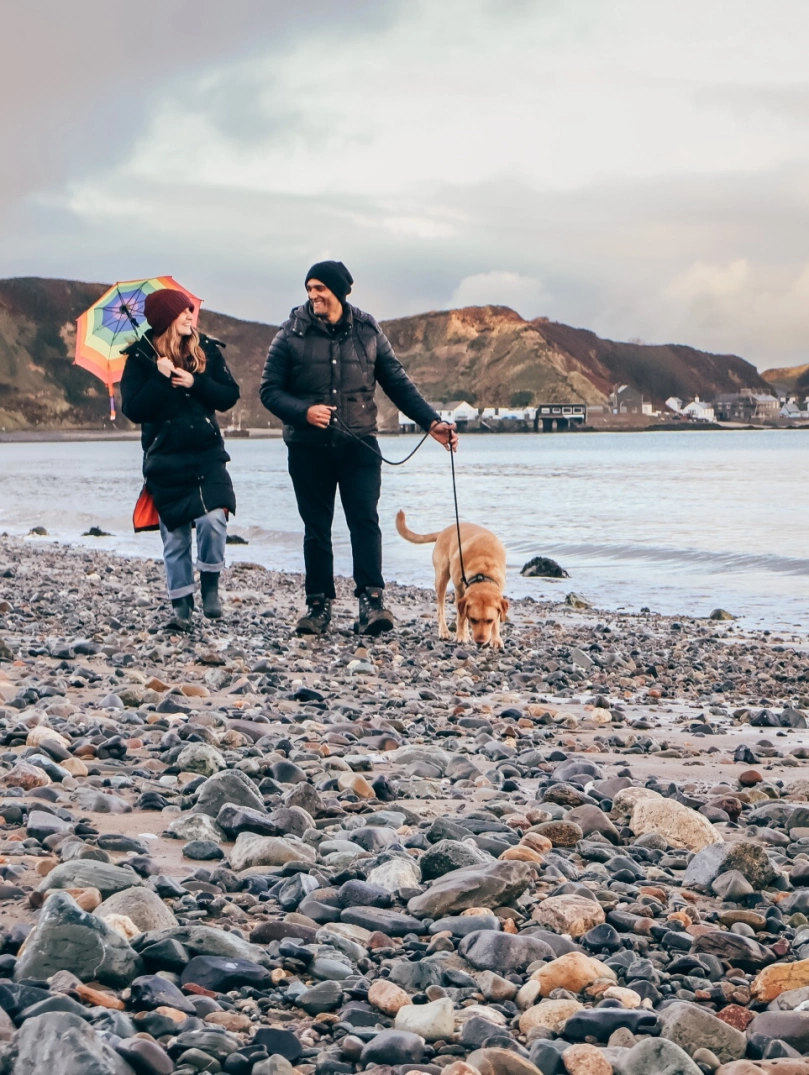 Cwpl a chi ar draeth cerrig mân wrth ymyl bae tawel, gyda phentref arfordirol pell a bryniau o dan awyr gymylog.