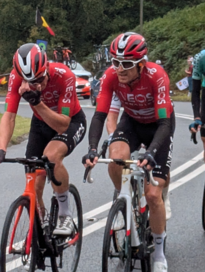 Grŵp o feicwyr ffordd proffesiynol mewn citiau rasio yn reidio'n agos gyda'i gilydd, gyda Geraint Thomas ar y blaen, ar ffordd wledig yn ystod digwyddiad beicio Tour of Britain 2025.