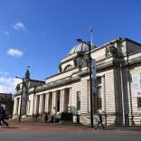 Tu allan i adeilad mawreddog gwyn Amgueddfa Genedlaethol Caerdydd ac awyr las yn gefndir.