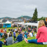 Pobl yn eistedd ar y gwair ar fryncyn yn edrych i lawr ar stondinau'r ŵyl fwyd