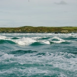 Llun o donnau yn y môr, a'r tir yn y cefndir