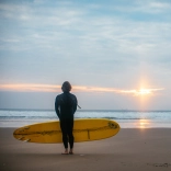 Dyn yn sefyll ar y traeth gyda'i gefn at y camera, yn wynebu'r môr ac yn dal bwrdd syrffio melyn