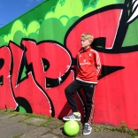 Bachgen yn pwyso ar wal gyda graffiti 'Cymru' arno, gydag un droed ar bêl-droed