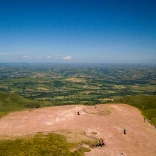 Golygfa o'r awyr o'r gwastadeddau gwyrdd islaw o ben Pen y Fan gydag awyr las.