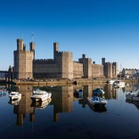 Llun o Gastell Caernarfon o'r de, gyda chychod yn yr harbwr o flaen y castell