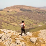 Menyw yn rhedeg ar draws rhostir gyda bryniau yn y cefndir. 