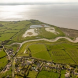 Golygfa o’r awyr o dirwedd werdd a dyfrffyrdd ger afon.