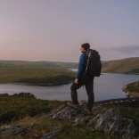 Dyn â bag ar ei gefn, yn sefyll ar ochr mynydd yn edrych allan ar draws tirwedd o fynyddoedd a llyn.