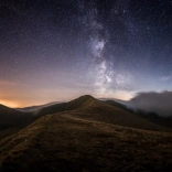 Awyr dywyll yn llawn sêr uwchben tirwedd fynyddig Bannau Brycheiniog gyda golau'r haul yn machlud ar y gorwel.